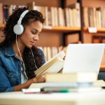 young female wearing headphones reading a book and studying on a laptop