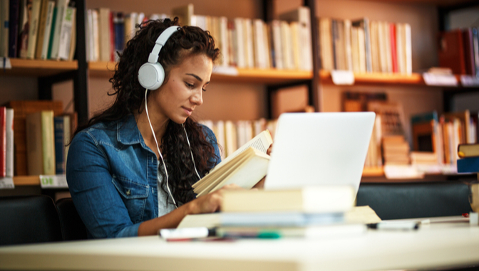 young female wearing headphones reading a book and studying on a laptop