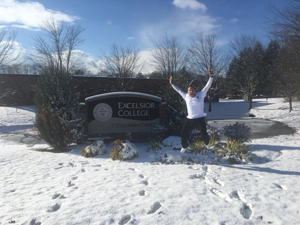 Andrew Lieberam jumping for joy in front of Excelsior University sign