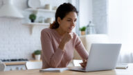 An adult student sits at home studying with her laptop and notebook.