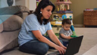 : A young mother sits at her kitchen table studying for class with her laptop while her daughter sits close by playing with a smartphone.