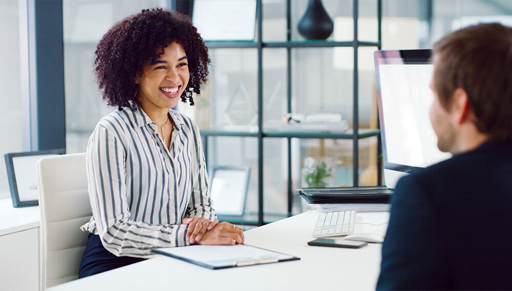 A hiring manager reaches across her desk shaking hands with a new employee as a smiling colleague looks on.