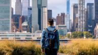 Man wearing a backpack gazes out on a path leading to a large city as he starts his journey