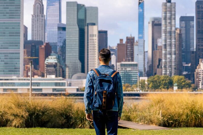 Man wearing a backpack gazes out on a path leading to a large city as he starts his journey