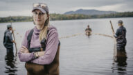 men and women standing in lake with waders on, woman in foreground looking at camera with arms folded