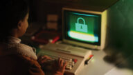 A woman in a turtleneck and a shearling coat types on the keyboard of a 1980s-era desktop computer with a green lock on the screen.