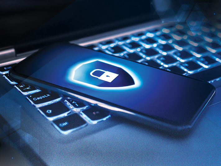 A smartphone with a glowing lock and shield on the screen rests on the backlit keyboard of a laptop.