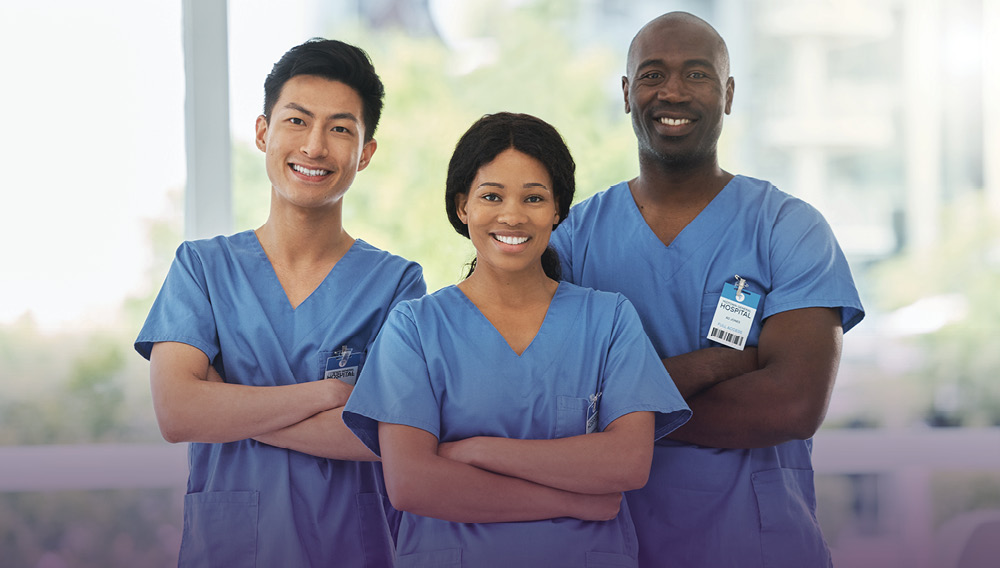 Three smiling nurses in blue scrubs standing with arms folded