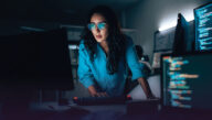 A woman in darkened room full of computer monitors leans over a keyboard and stares at a screen.