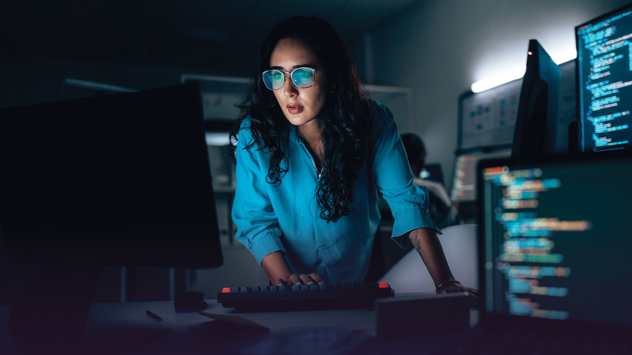 A woman in darkened room full of computer monitors leans over a keyboard and stares at a screen.