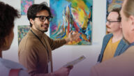 An art professor gestures toward a painting in a museum as he discusses it with the students around him.