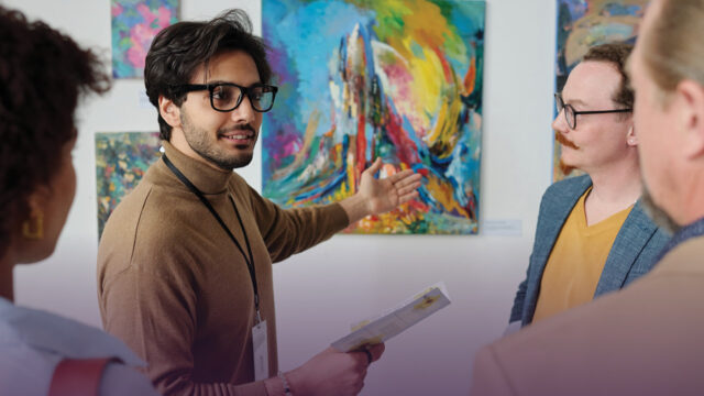 An art professor gestures toward a painting in a museum as he discusses it with the students around him.