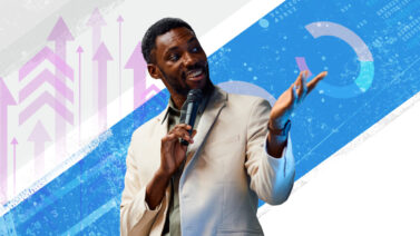 A man in a sport coat holds a microphone in one hand and gestures toward a presentation with the other.