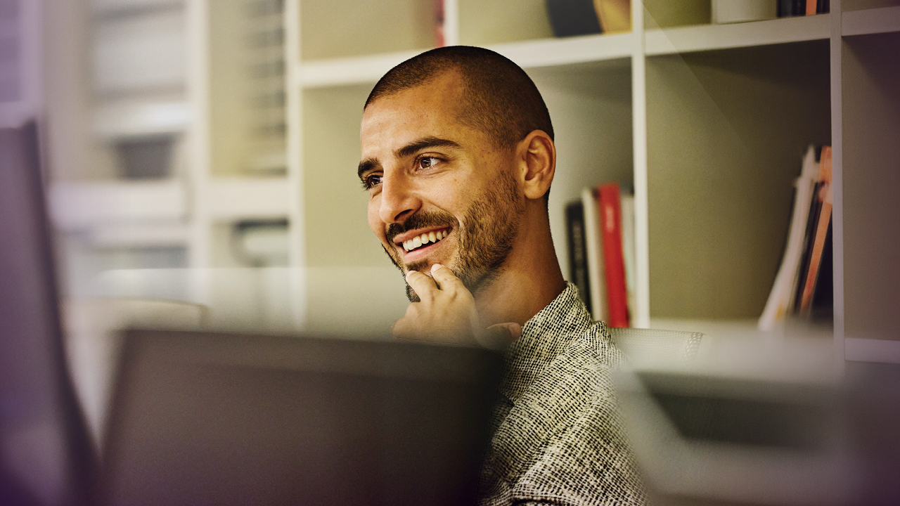 An MBA student smiles and touches his chin as he looks at his computer screen.
