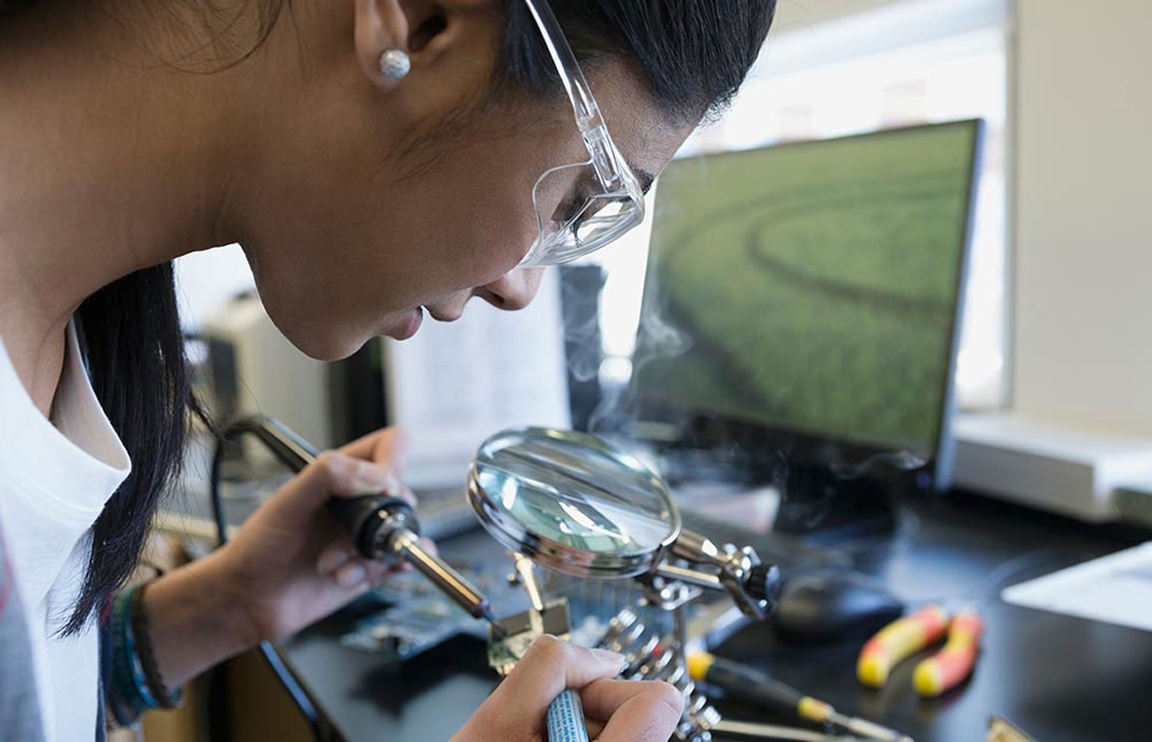 A student wearing safety glasses uses a magnifying lamp to closely inspect an electronic circuit board.