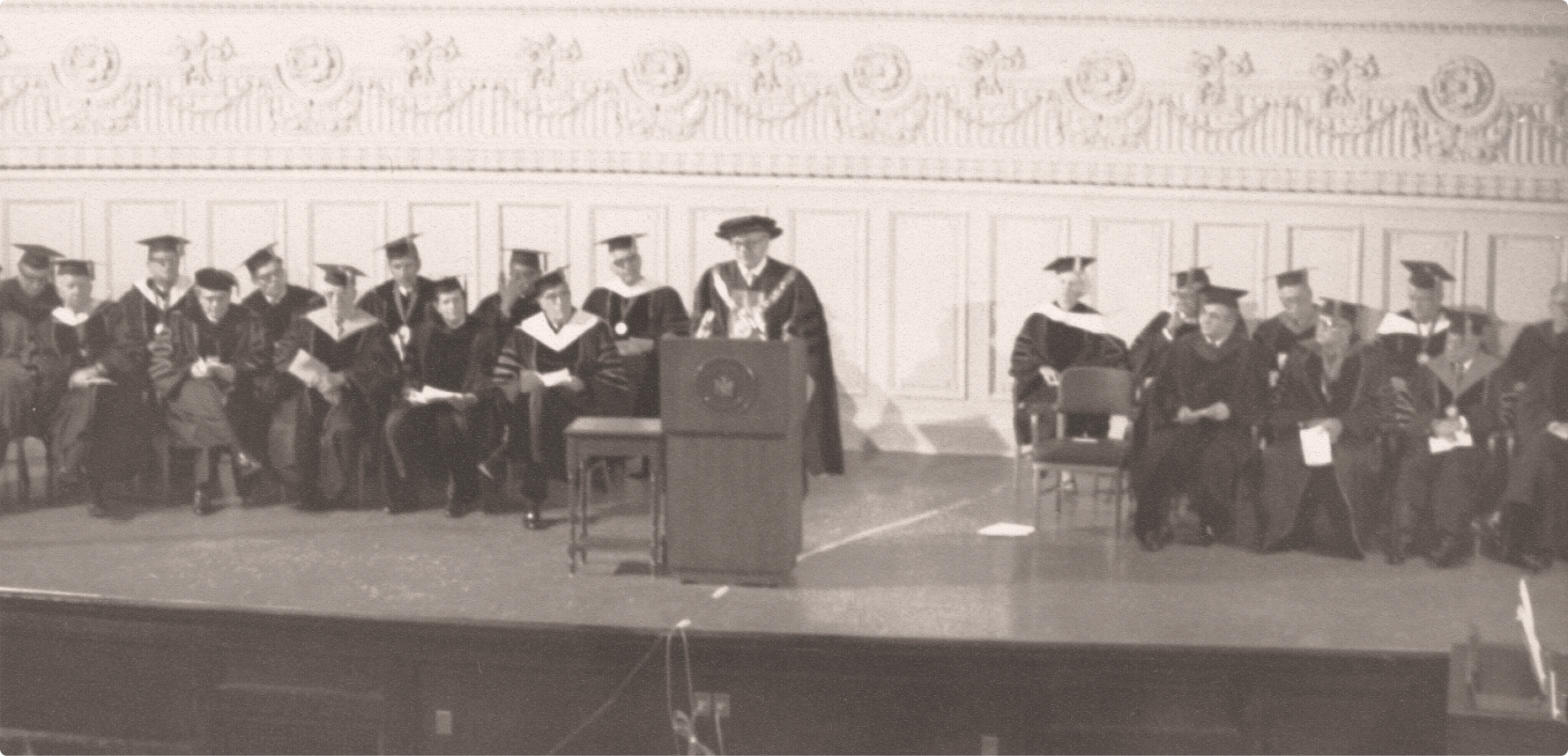 Faculty and graduates in academic regalia gather on stage during an early Excelsior University commencement ceremony.